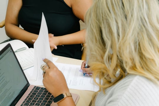 Twee collega’s bekijken samen documenten op een laptop en op papier
